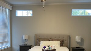 A bedroom with a bed, a ceiling fan, two bedside tables with lamps, a window, and a nightstand with a basket of toiletries.