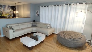 A modern living room with a sectional sofa, a glass coffee table, a round ottoman, and a painting on the wall.