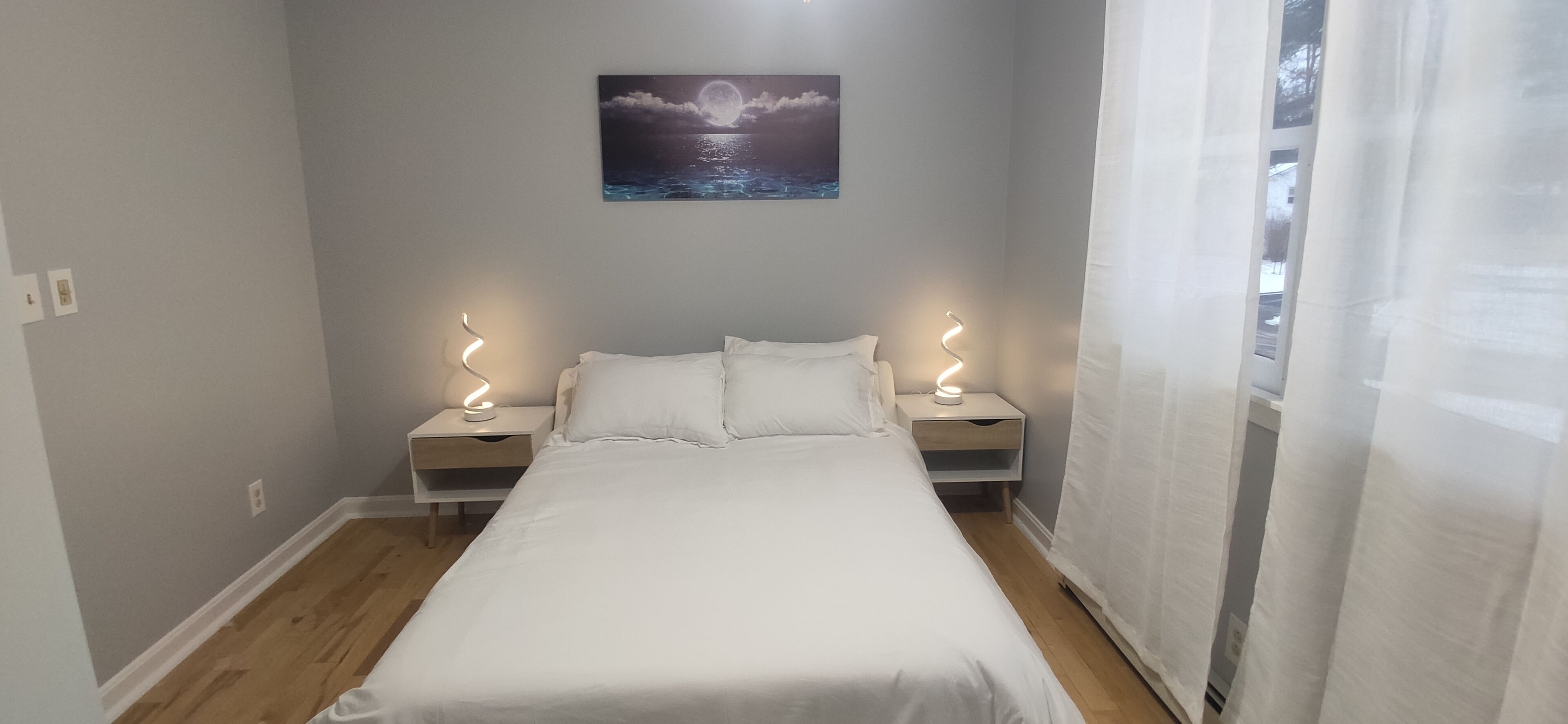 A neatly made bed in a room with light grey walls, a painting of a moon over water, and two bedside lamps.