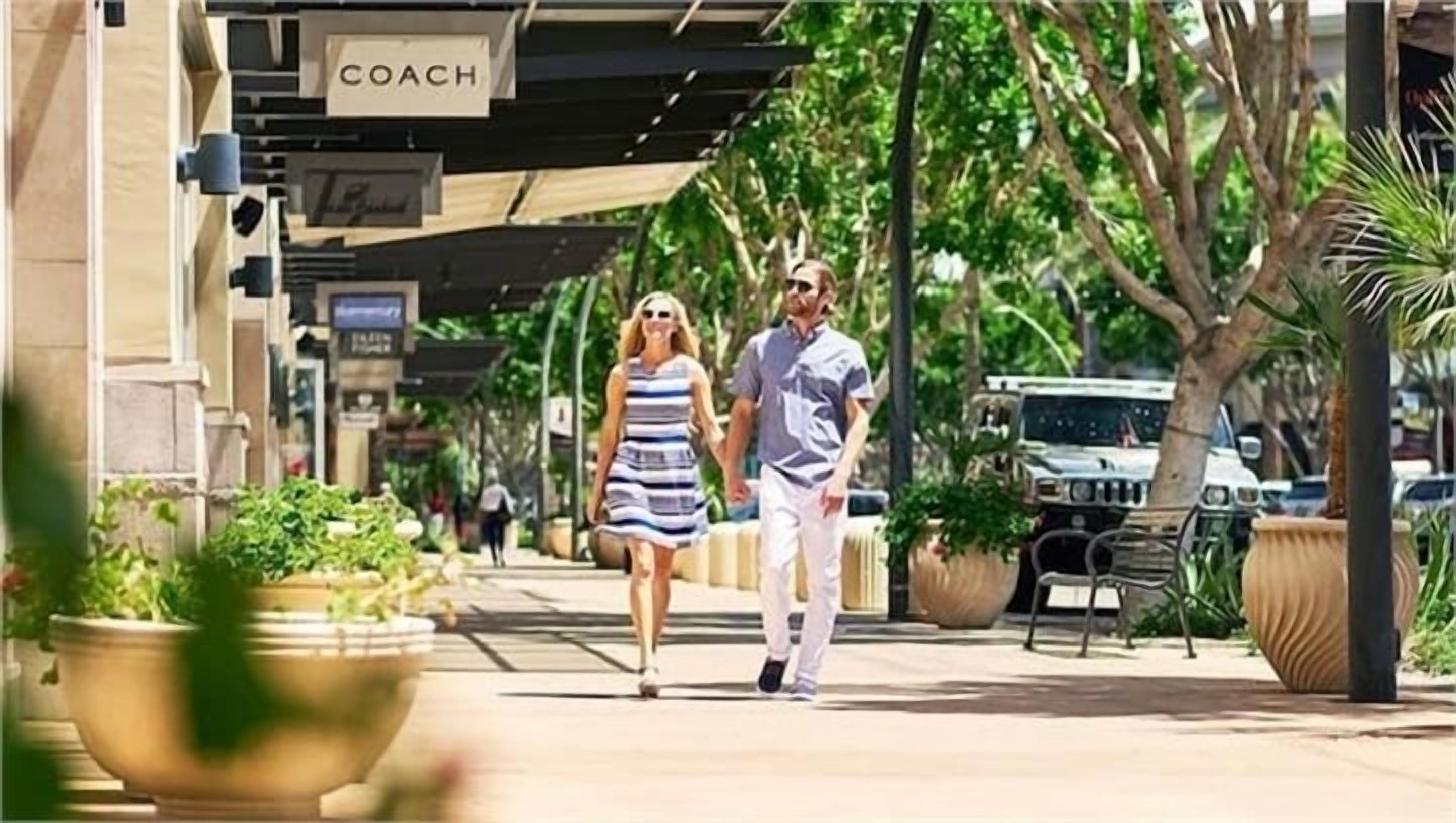Un couple se promène sur un trottoir bordé de jardinières, devant un bâtiment sur lequel on peut lire l'inscription « COACH ».