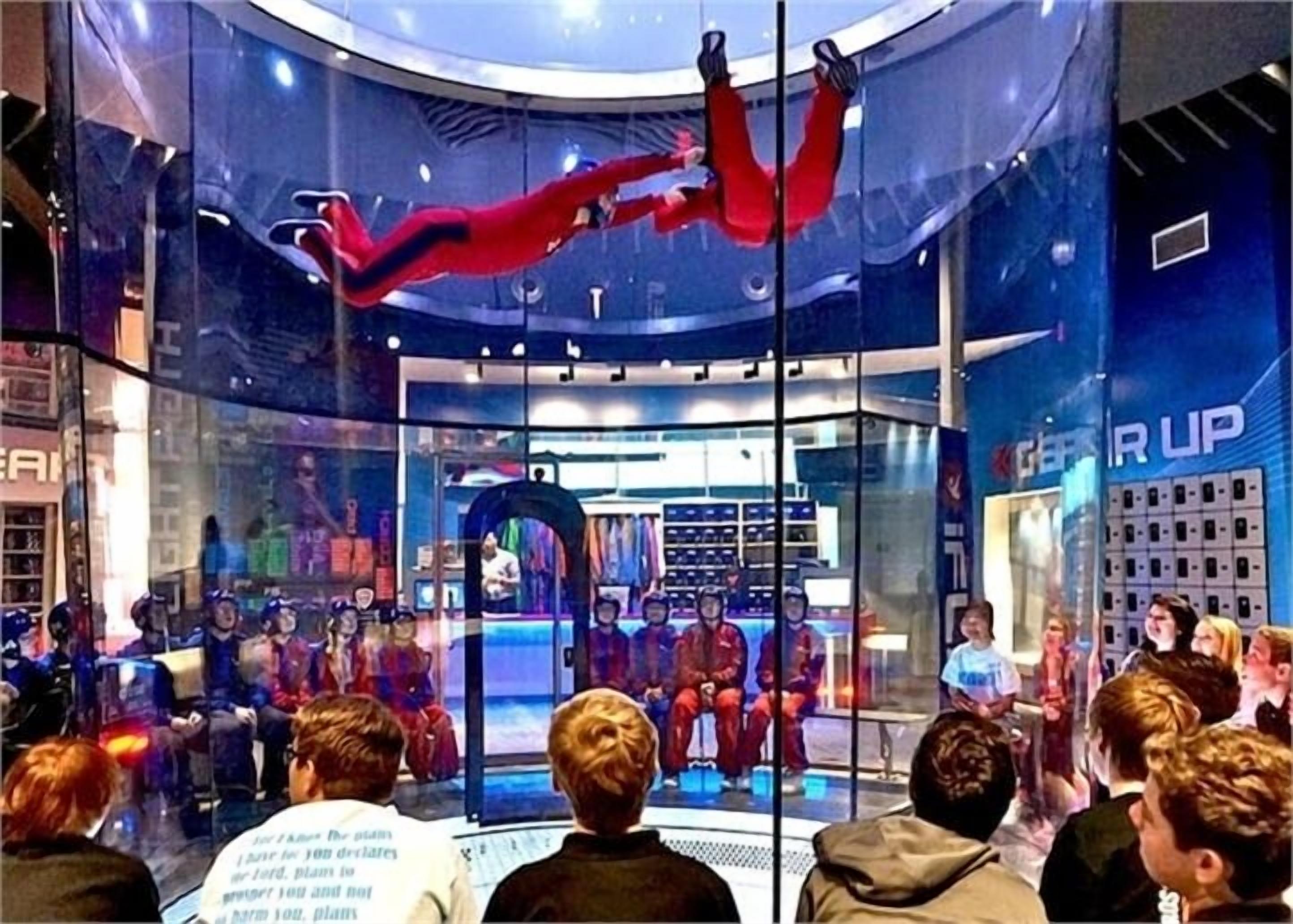 A glass-walled room with a circular ceiling, a group of people seated facing a stage, and a person performing an act.