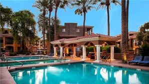 A swimming pool with a clear blue water, surrounded by palm trees and a building with a tiled roof. - 7 Bedroom / 6 Bath Condos at TPC Scottsdale (Scottsdale)