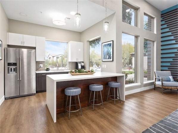 A modern kitchen with a central island, stainless steel appliances, and large windows offering views of the outdoors.