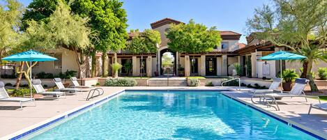 A swimming pool with lounge chairs and umbrellas, surrounded by trees and a building with a tiled roof.