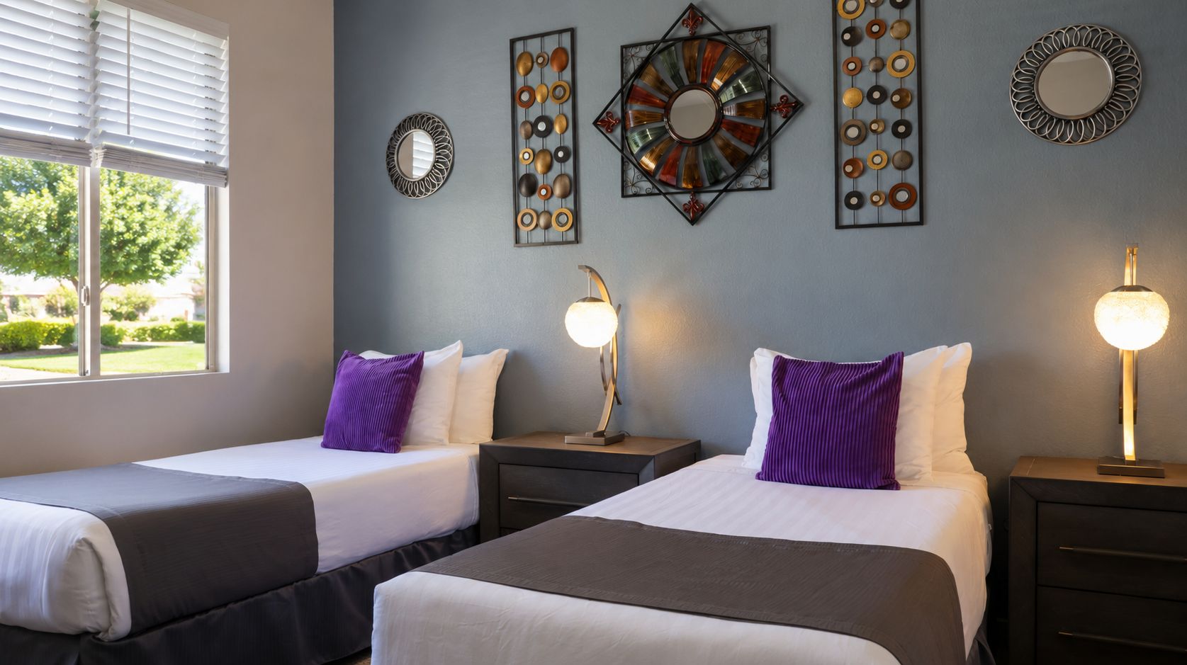 A hotel room with two beds, a window with blinds, decorative wall art, and bedside lamps.