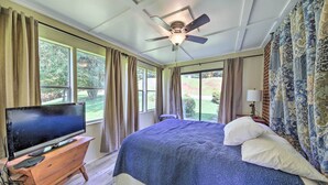 Un dormitorio con cama, televisor sobre una cómoda de madera, ventilador de techo y ventanas amplias con cortinas.