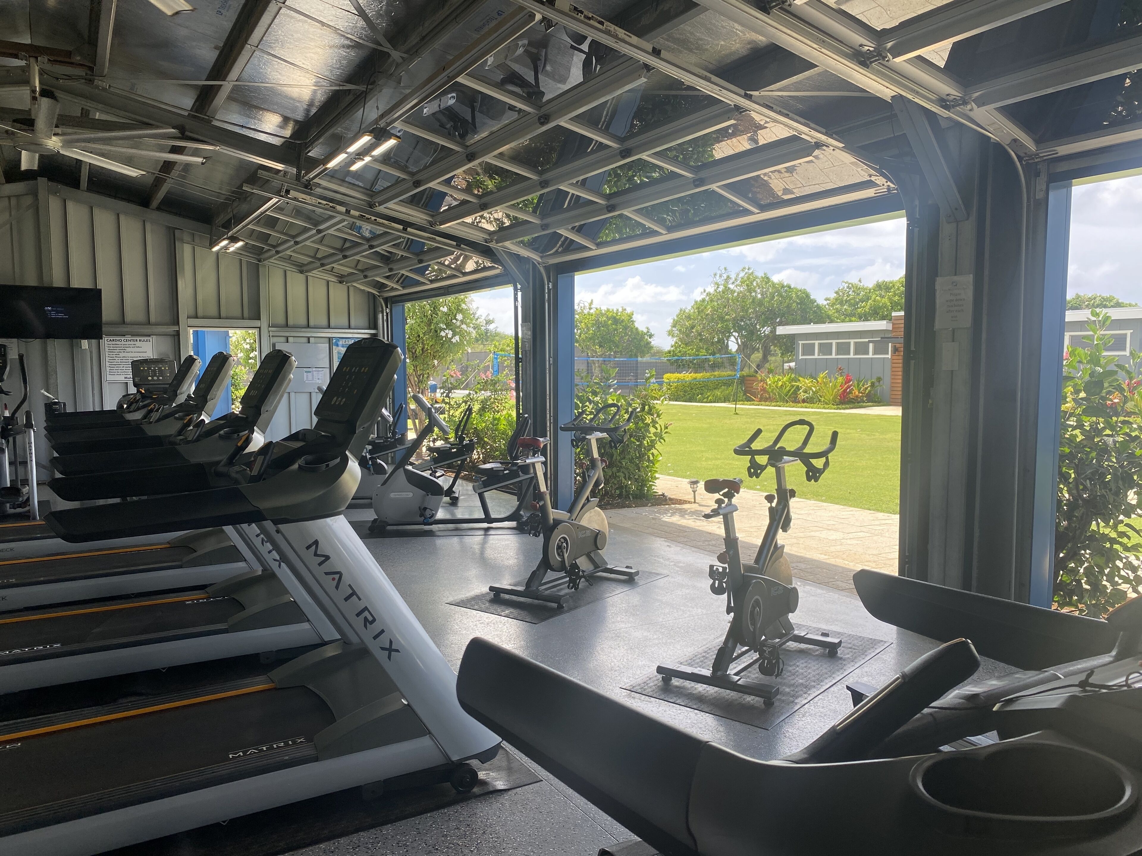 A gym with cardio equipment, including treadmills and elliptical machines, and a view of a well-maintained outdoor area with grass and trees.