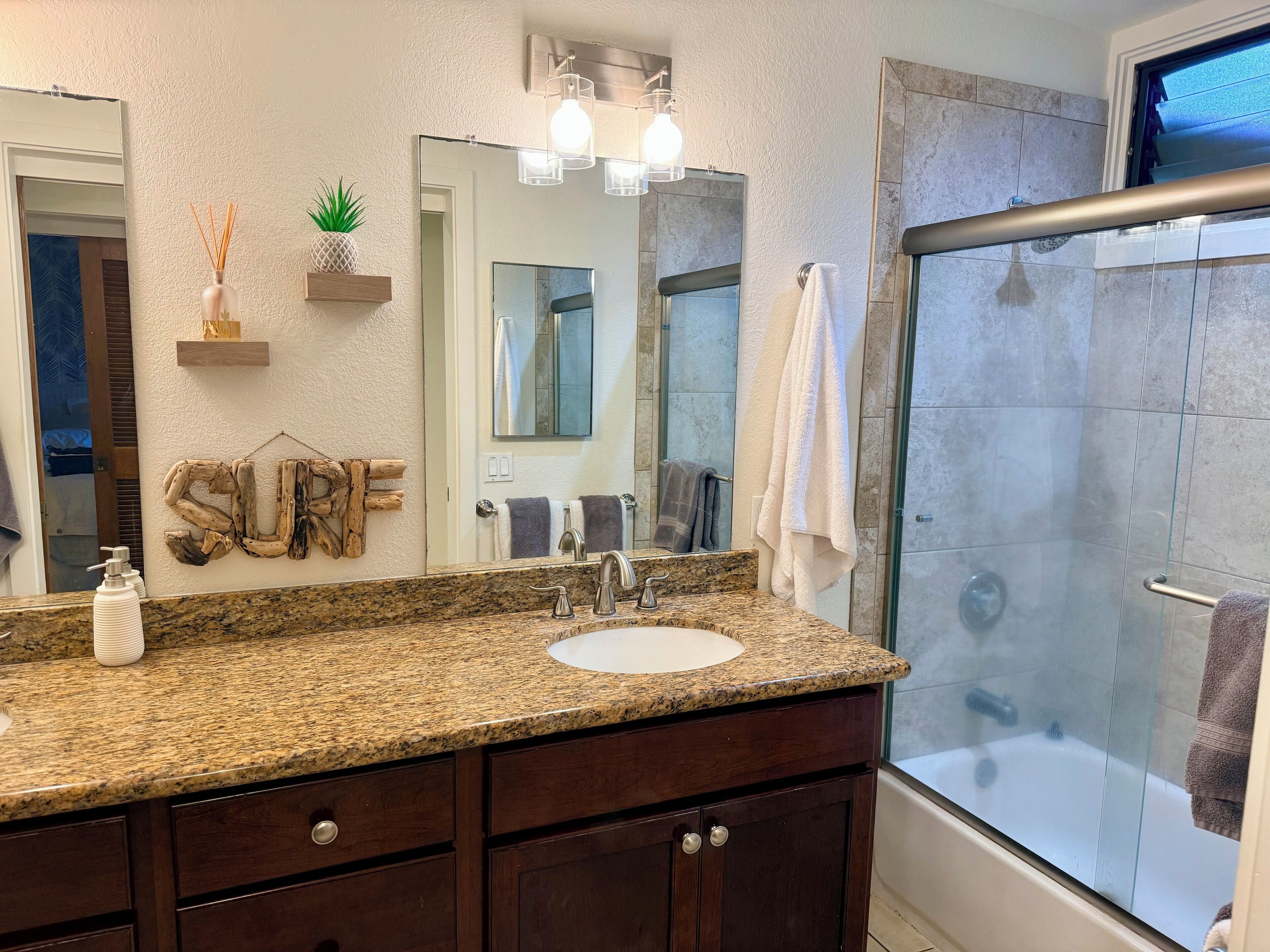 Une salle de bain dotée d’un comptoir en granit, d’un lavabo blanc, d’un grand miroir, d’une douche avec des portes en verre et d’un porte-serviettes.