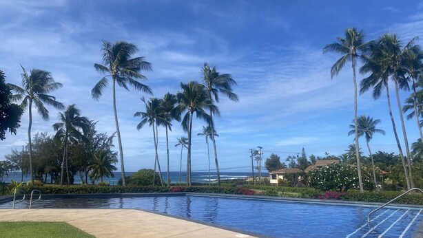 A swimming pool with a clear view of the ocean, surrounded by palm trees and a well-maintained lawn. - Poipu Paradise Beach Condo (Koloa)