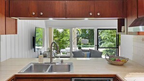 A modern kitchen with wooden cabinets, a stainless steel sink, and a dishwasher. There is a window with a view of greenery outside. - Poipu Paradise Beach Condo (Koloa)