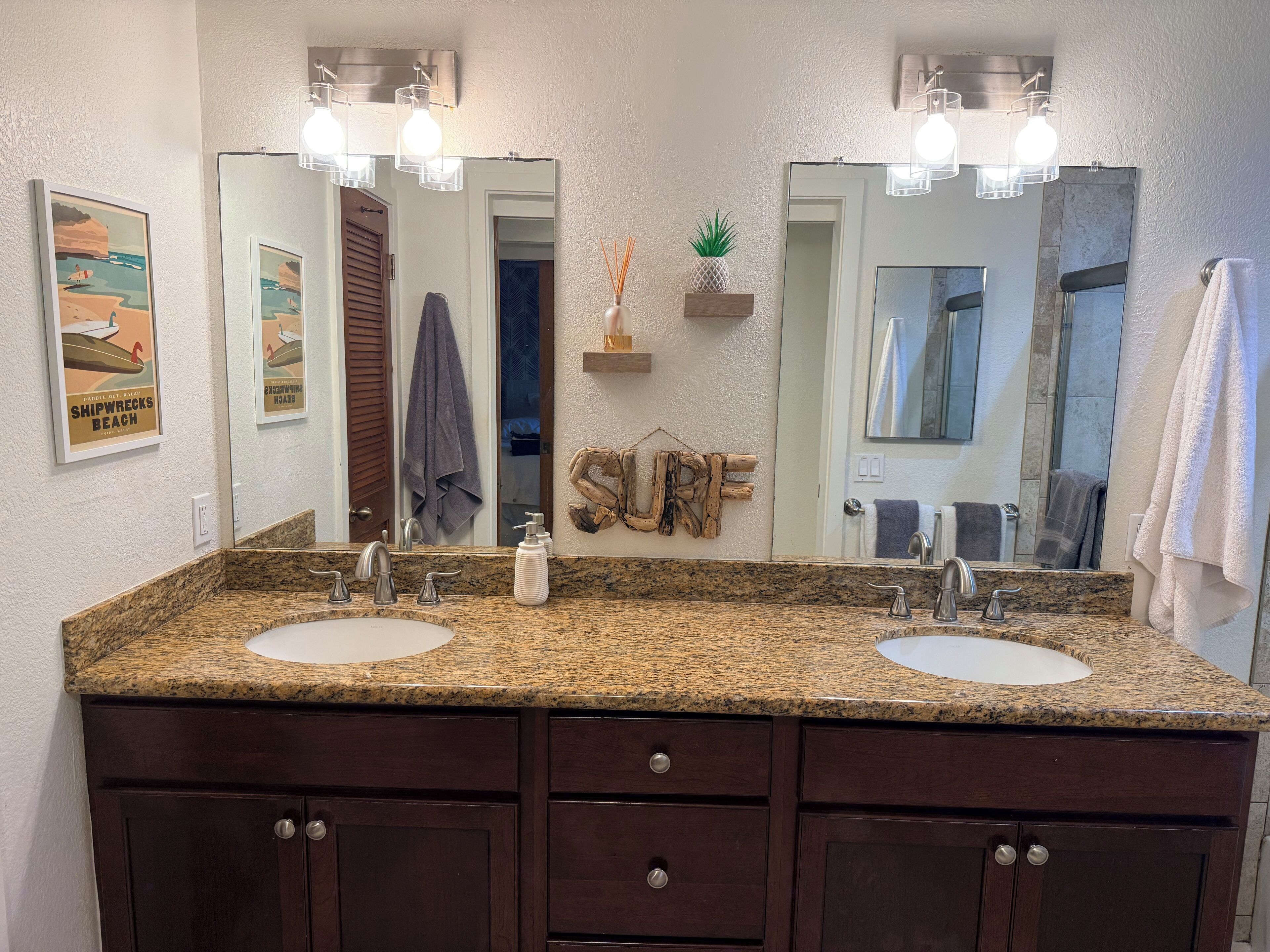 A bathroom with two sinks, dark wooden cabinets, a mirror with lights, a towel rack, and a 'SURF' sign.