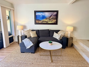 A modern living room with a dark grey sectional sofa, two side tables with lamps, a framed landscape painting, and a small round table with a plant.