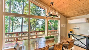 A dining area with a large wooden table, four chairs, and a chandelier. There are large windows with a view of trees and a deck.
