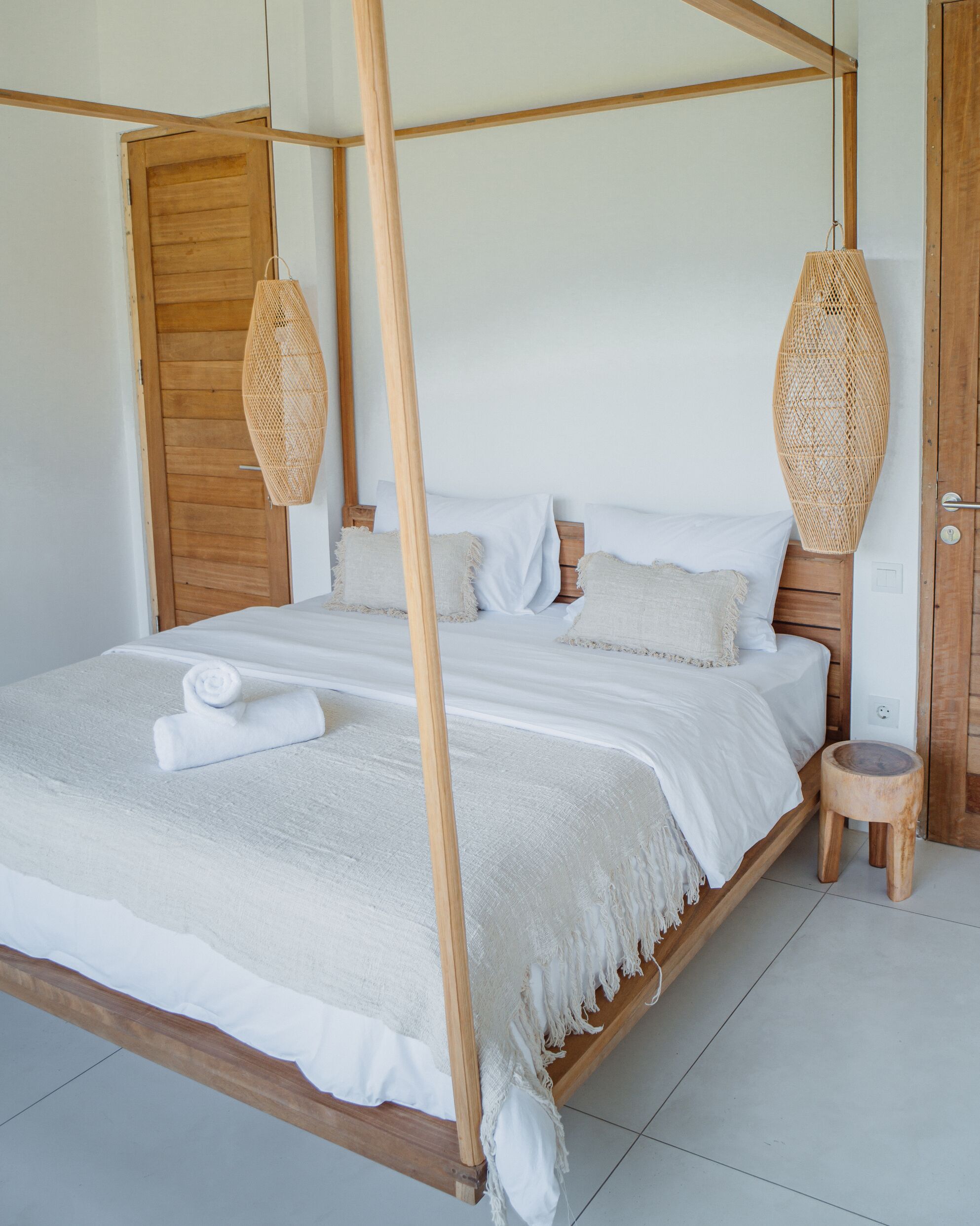 Uma cama de dossel com estrutura de madeira e roupa de cama branca, uma porta de madeira e um banquinho de madeira.
