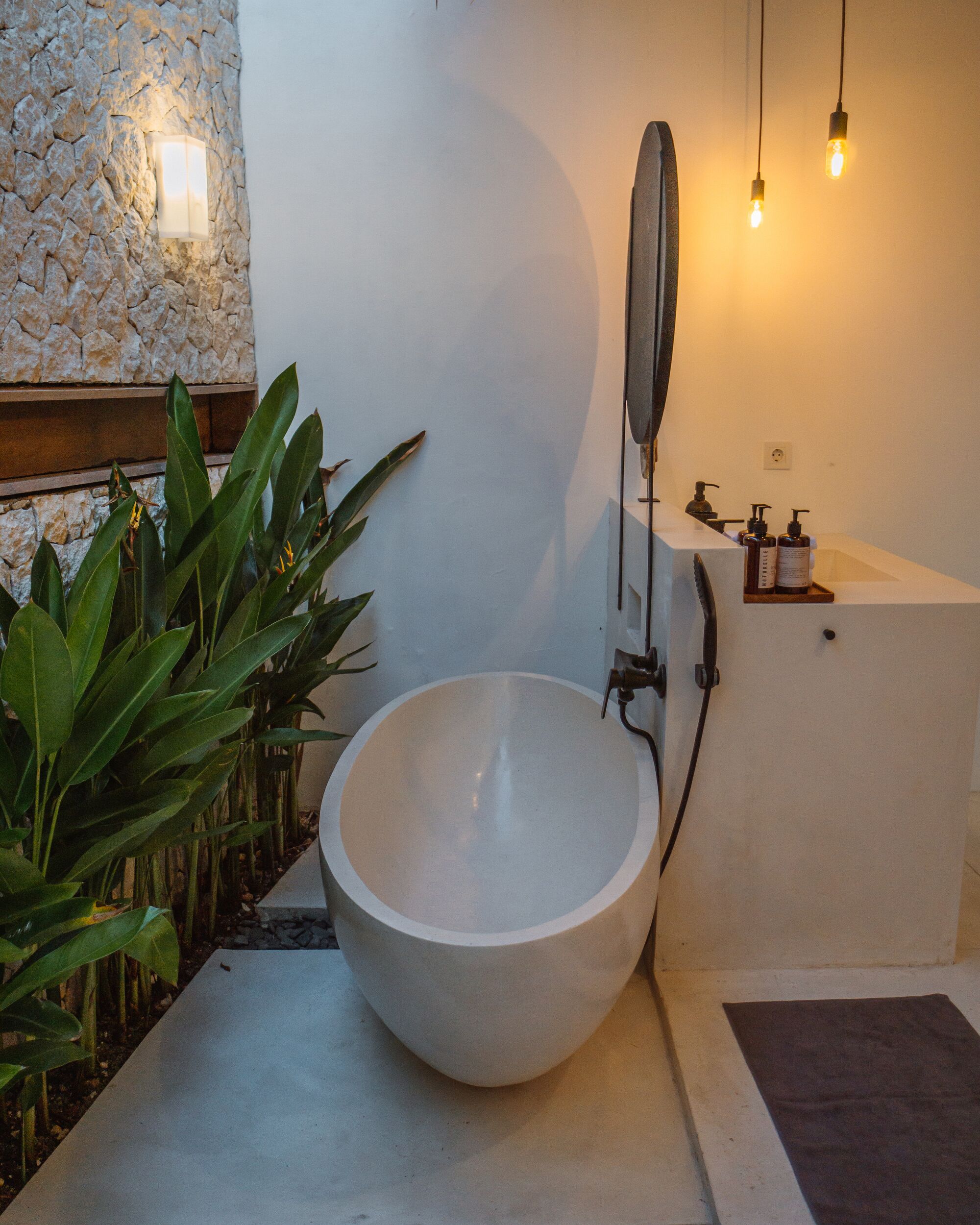 Une salle de bain avec un grand lavabo rond, un miroir et une baignoire. Des plantes se trouvent sur le côté gauche et un mur de pierre en arrière-plan.