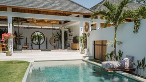 Un espace piscine avec une eau d’un bleu limpide, un coin salon couvert agrémenté de coussins et un palmier.