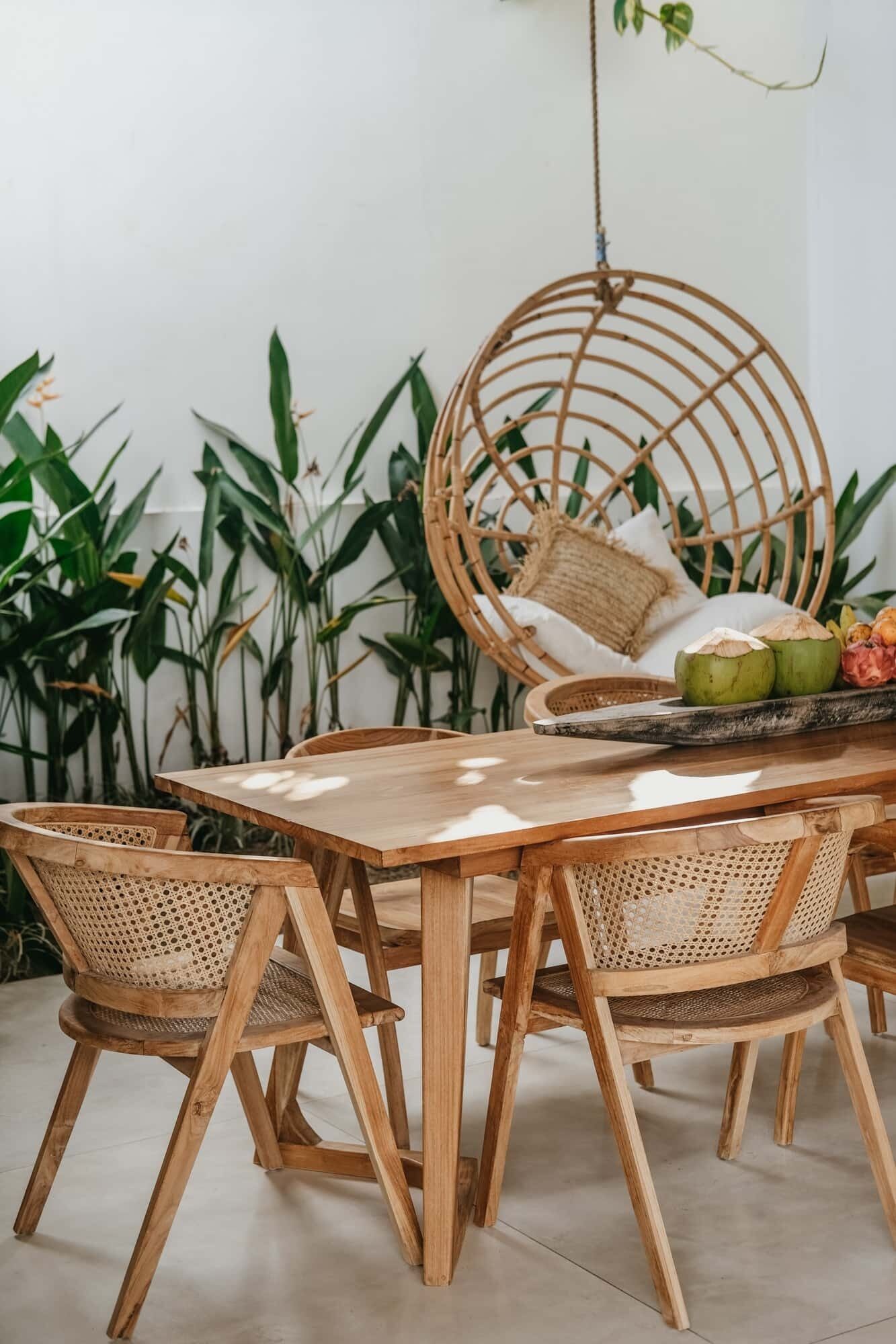 A wooden dining table with four woven chairs, a hanging rattan chair, and a tray with coconuts and fruits.