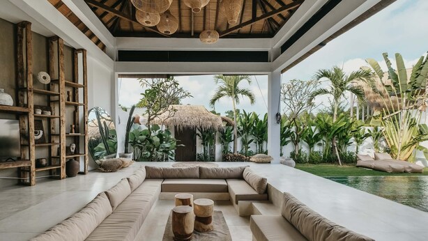 Un intérieur moderne doté d’un grand espace salon, d’étagères en bois et offrant une vue sur des plantes tropicales et une piscine.