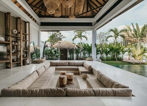 A modern interior with a large seating area, wooden shelves, and a view of tropical plants and a pool.