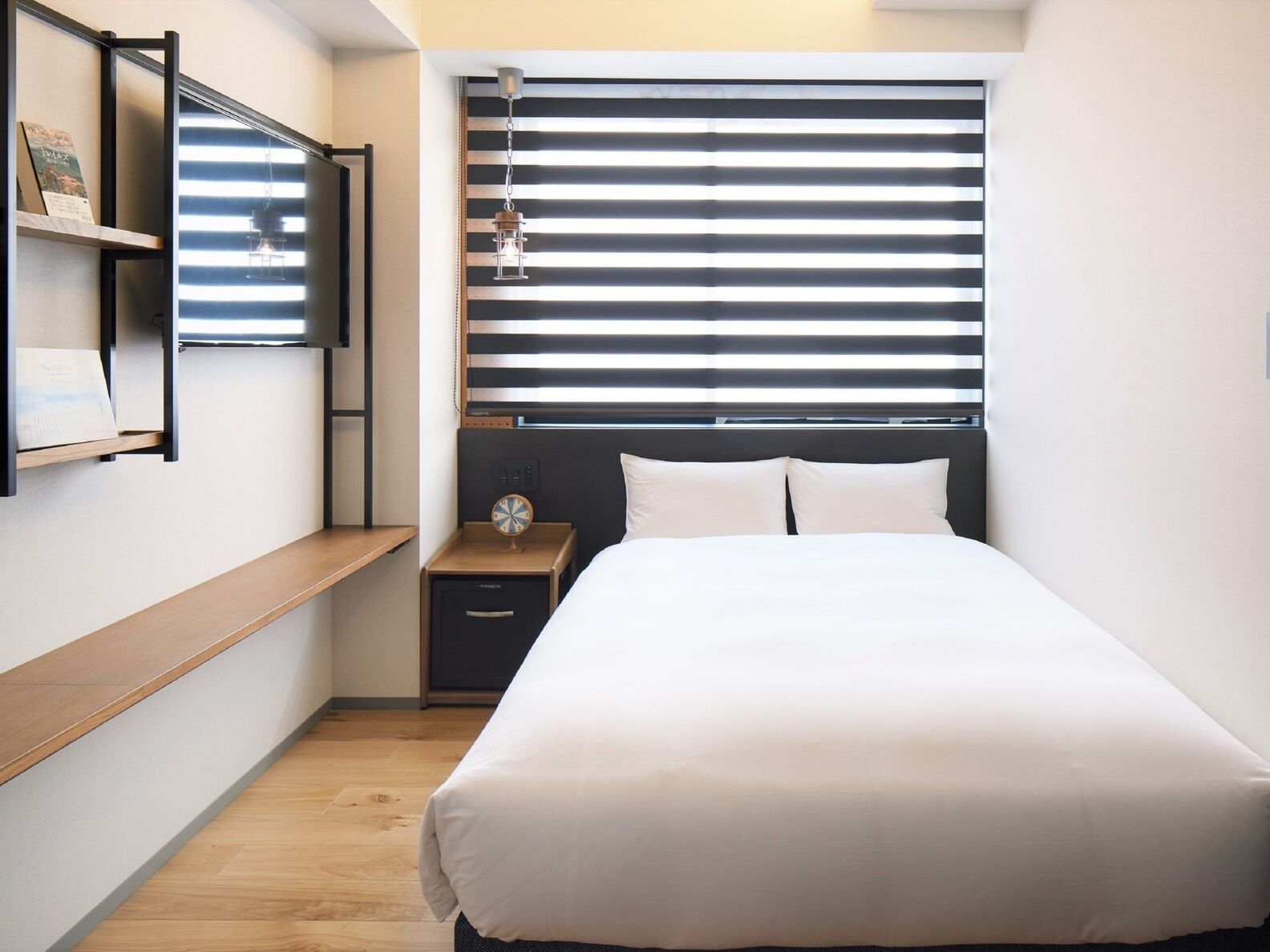 A bedroom with a bed, a nightstand, a shelf with a clock, and a window with blinds.