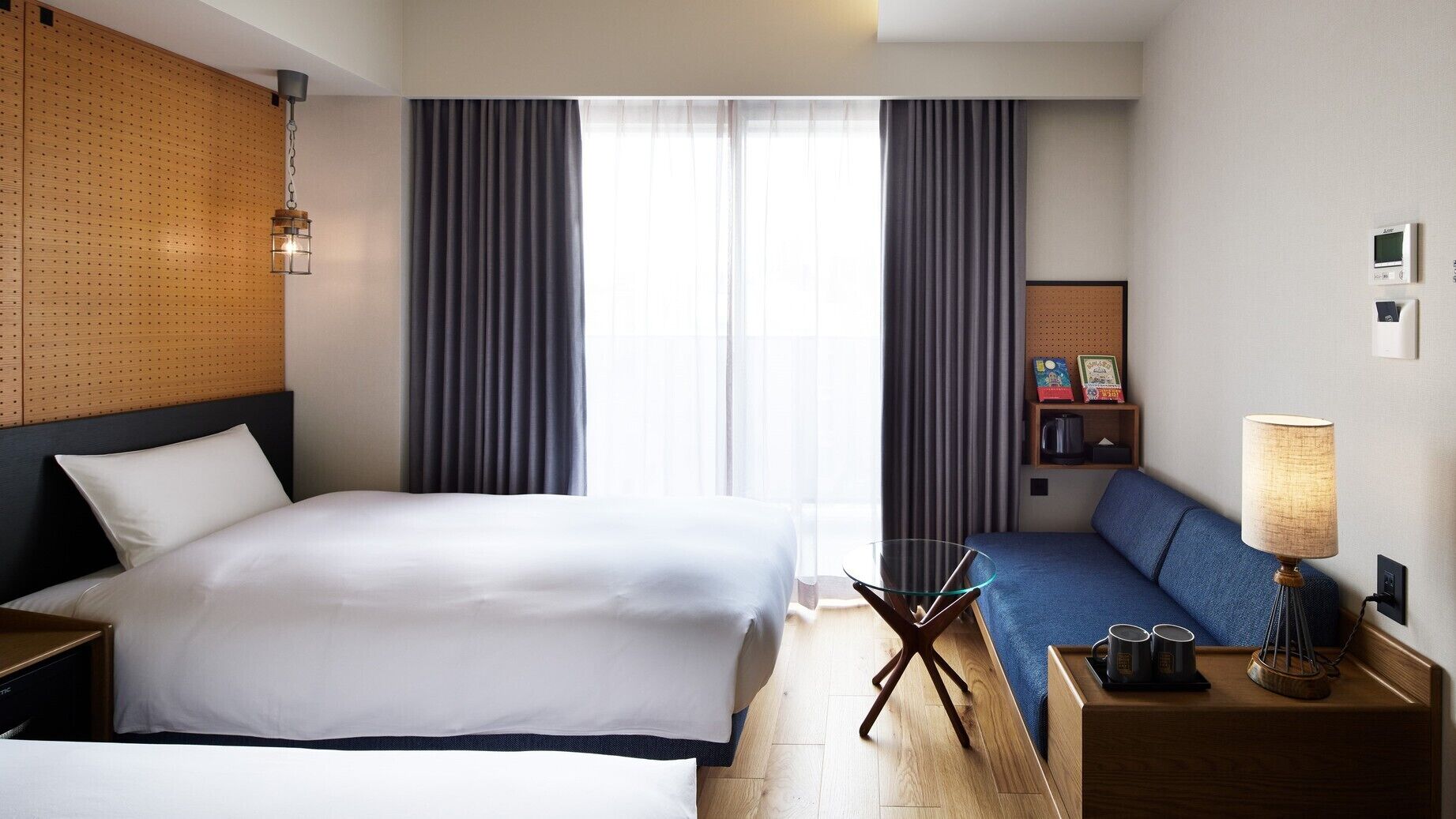 A hotel room with a large bed, a sofa, a desk with a lamp, a coffee machine, and a wall-mounted thermostat.
