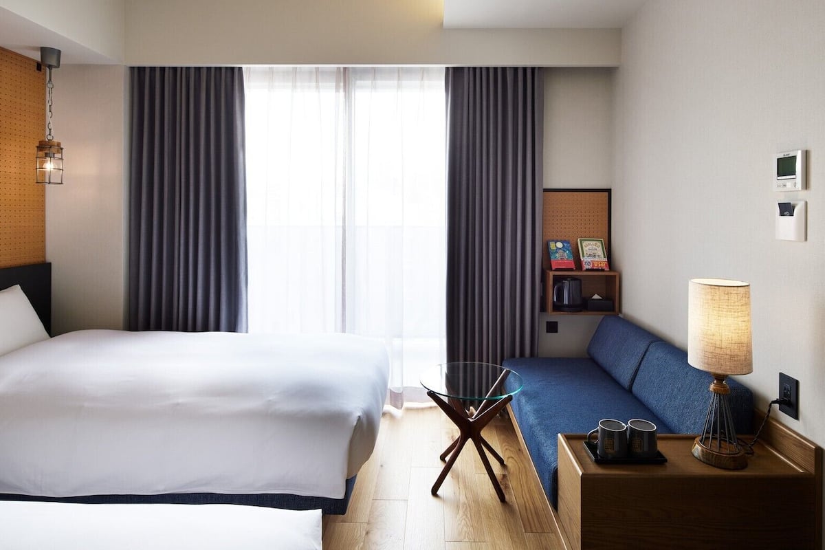 Habitación de hotel con una cama grande, un sofá, un escritorio con lámpara, una cafetera y un termostato fijado a la pared.
