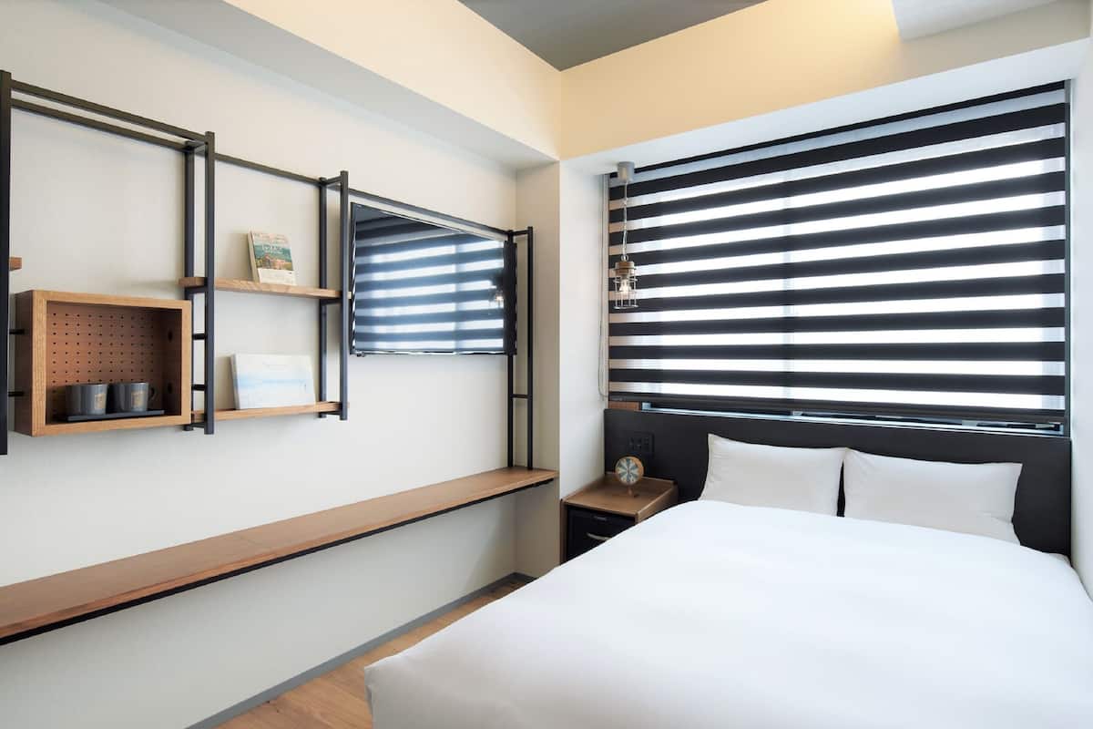 Una habitación de hotel moderna con una cama grande, un banco de madera, una mesita con un reloj y una ventana con persianas horizontales.