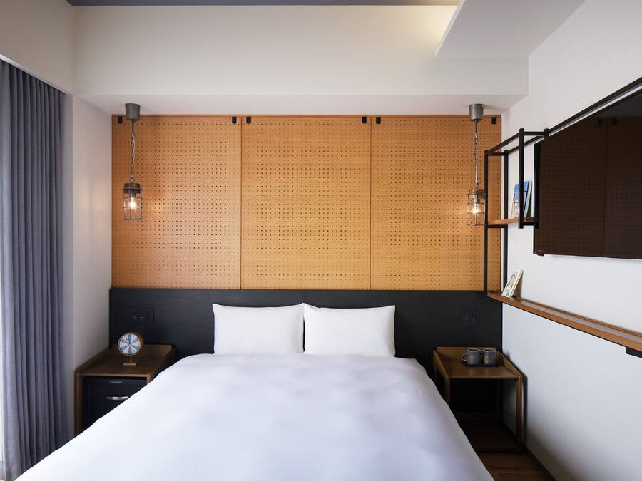 A modern hotel room with a large bed, wooden headboard, two bedside tables, a nightstand, a wall-mounted TV, and a window with curtains.