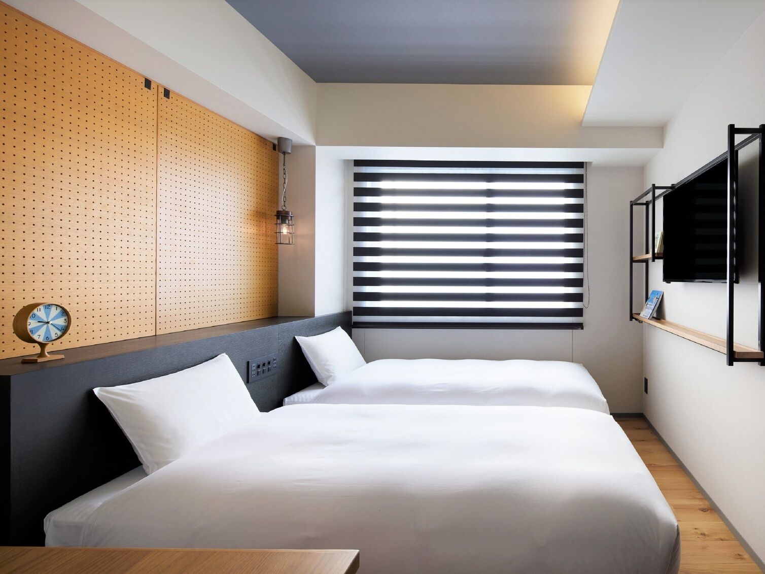 A modern hotel room with a bed, a wall clock, a window with blinds, and a wooden accent wall.
