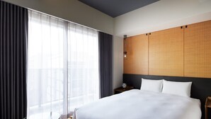 Une chambre d’hôtel avec un grand lit, une table en verre, une chaise, une table de chevet et une vue sur la ville à travers de fines rideaux.