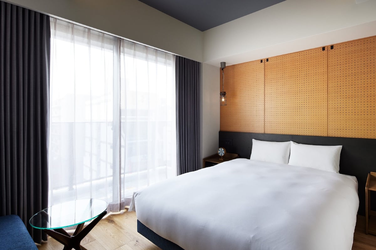 Habitación de hotel con una cama grande, una mesita de vidrio, una silla, una mesita de noche y vistas a la ciudad a través de cortinas traslúcidas.