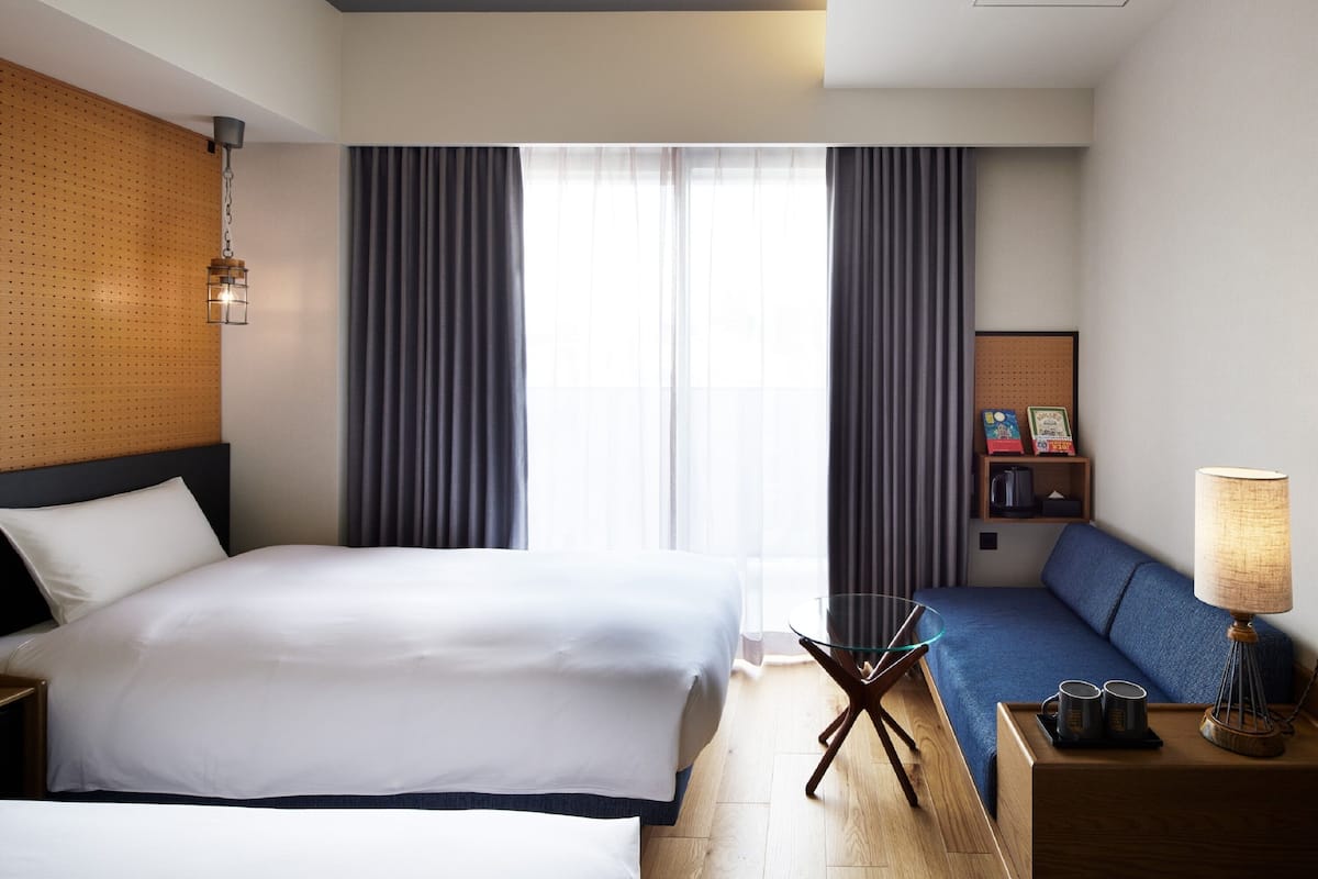 Una habitación de hotel con una cama grande, un sofá más pequeño, una mesa de centro de vidrio, una lámpara y un estante de pared con objetos decorativos.