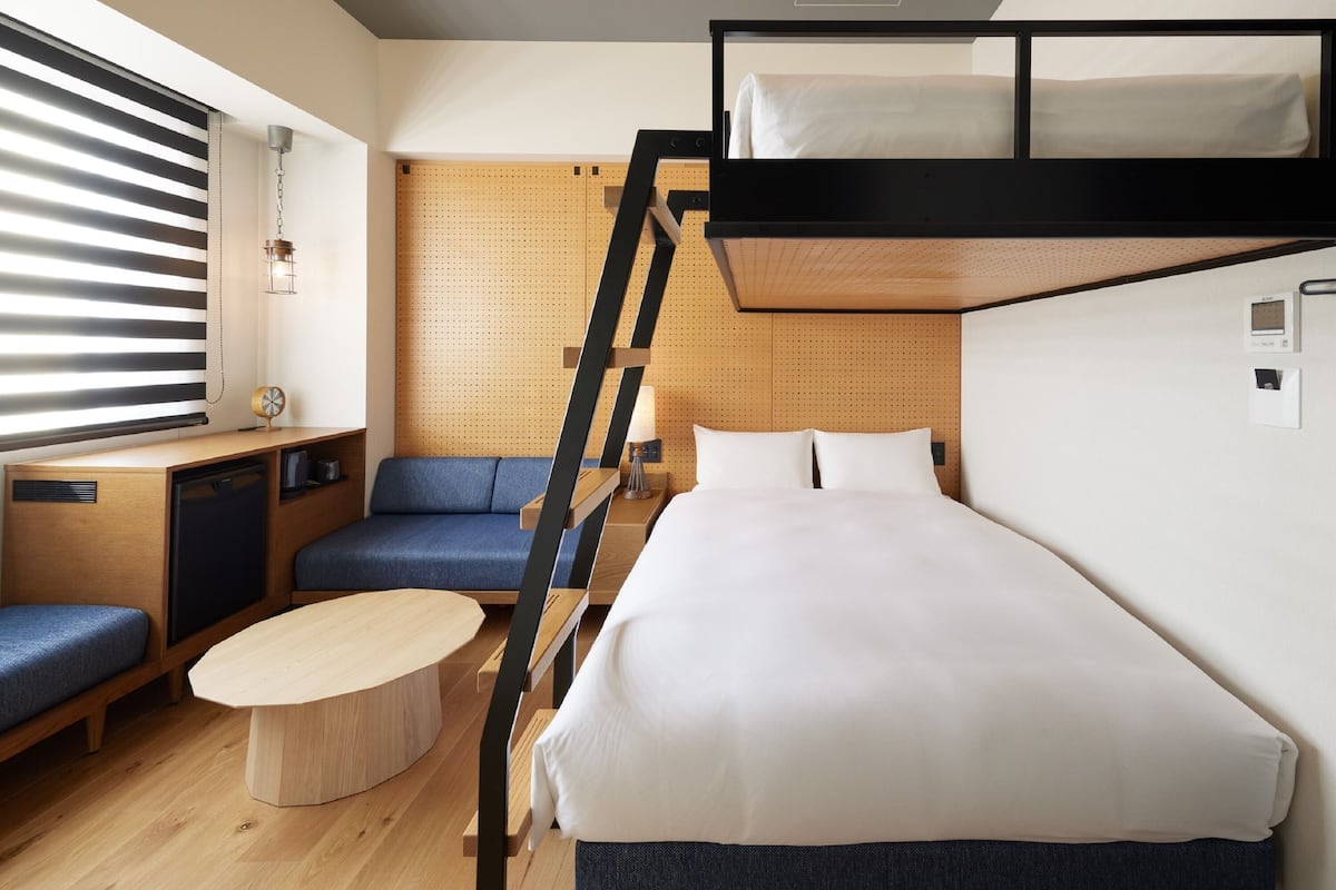 Habitación de hotel compacta con literas, un escritorio con silla, una mesita redonda pequeña y un sofá.