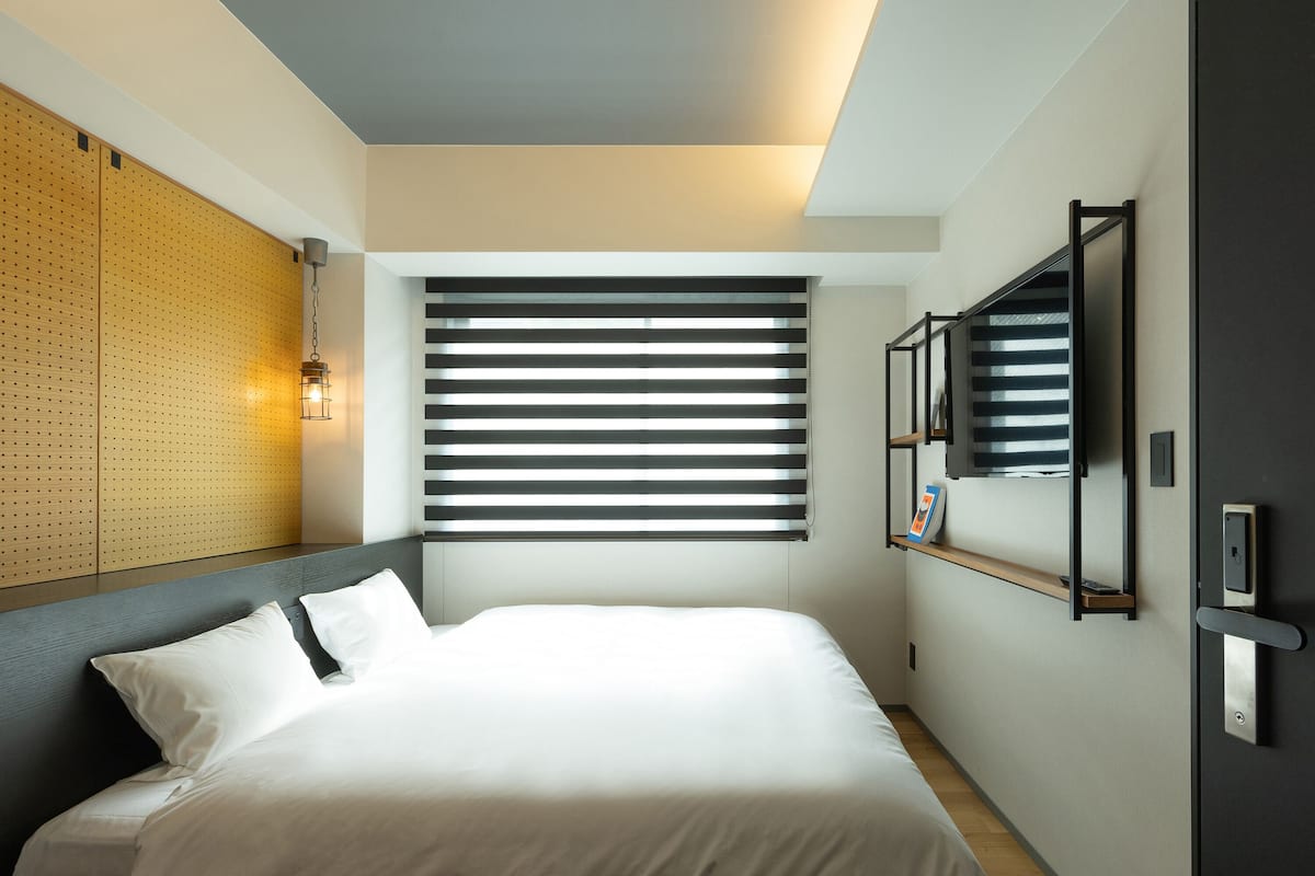 Una habitación de hotel moderna con una cama grande, un cabecero de madera, un televisor montado en la pared y una ventana con persianas.