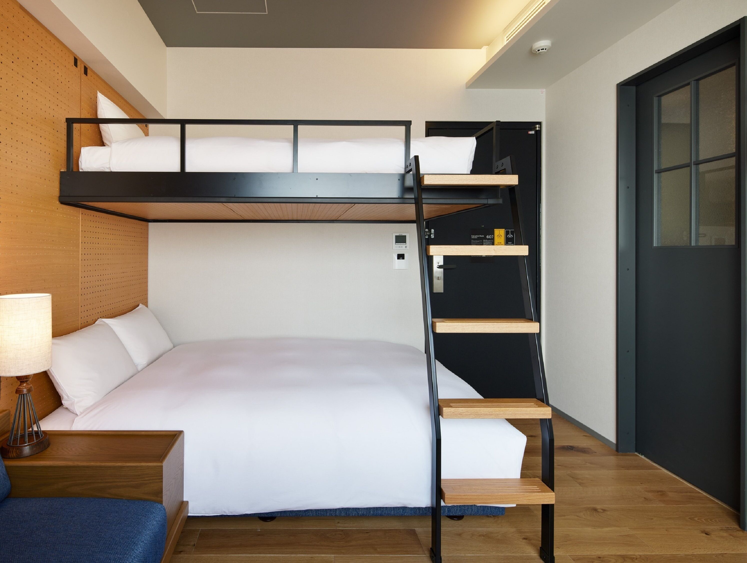 A bunk bed with a wooden ladder, a bedside table with a lamp, and a door with a window.