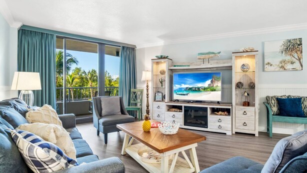 A modern living room with a large TV, comfortable seating, and a balcony with curtains. - Beautifully Updated Beachfront Condo w/Resort Amenities, including Heated Pool & WiFi (Marco Island)