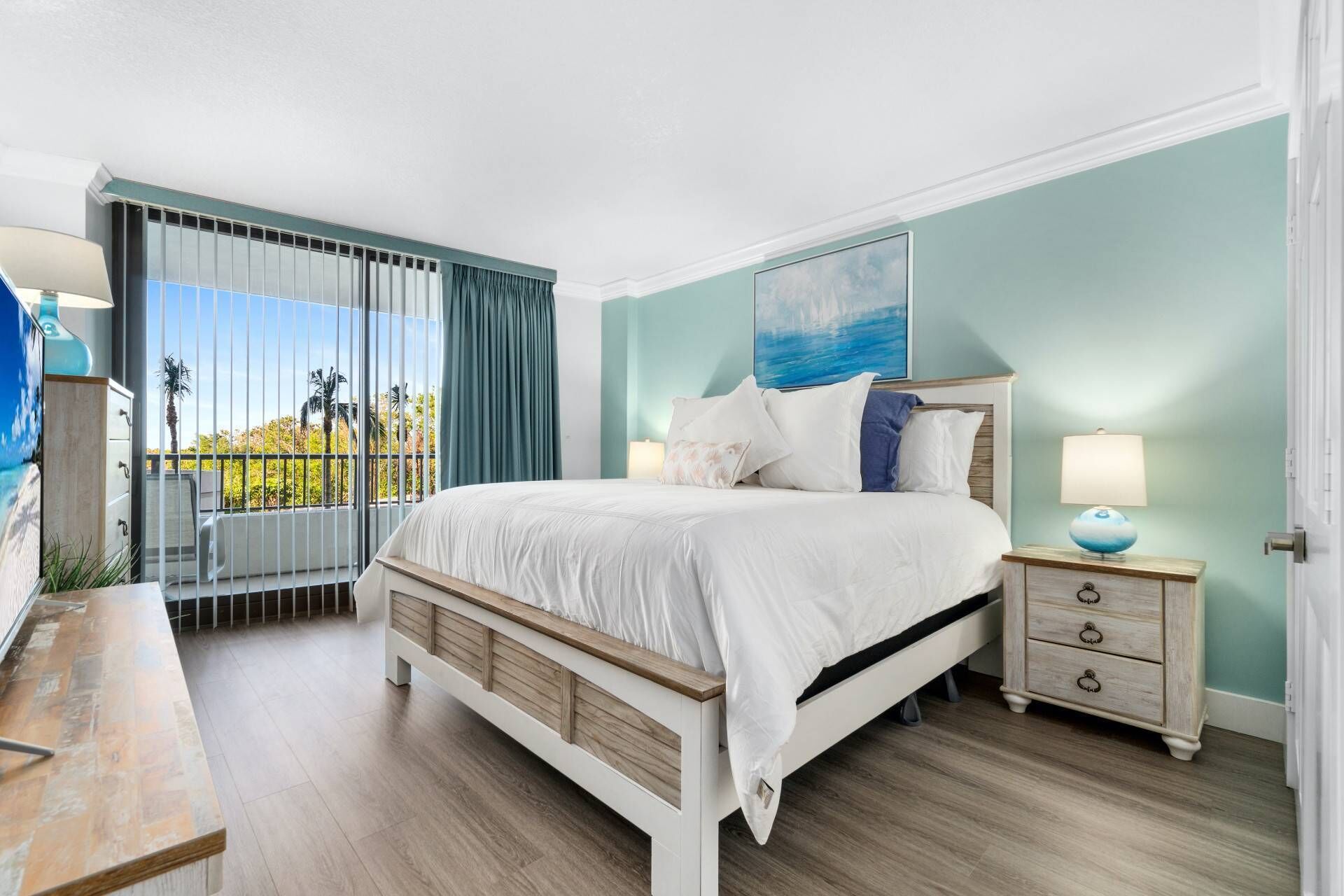 A bedroom with a bed, bedside tables, a nightstand, a window with curtains, and a view of palm trees.