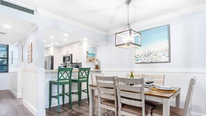 A dining area with a wooden table and chairs, a kitchen in the background, and a beach-themed painting on the wall. - Beautifully Updated Beachfront Condo w/Resort Amenities, including Heated Pool & WiFi (Marco Island)