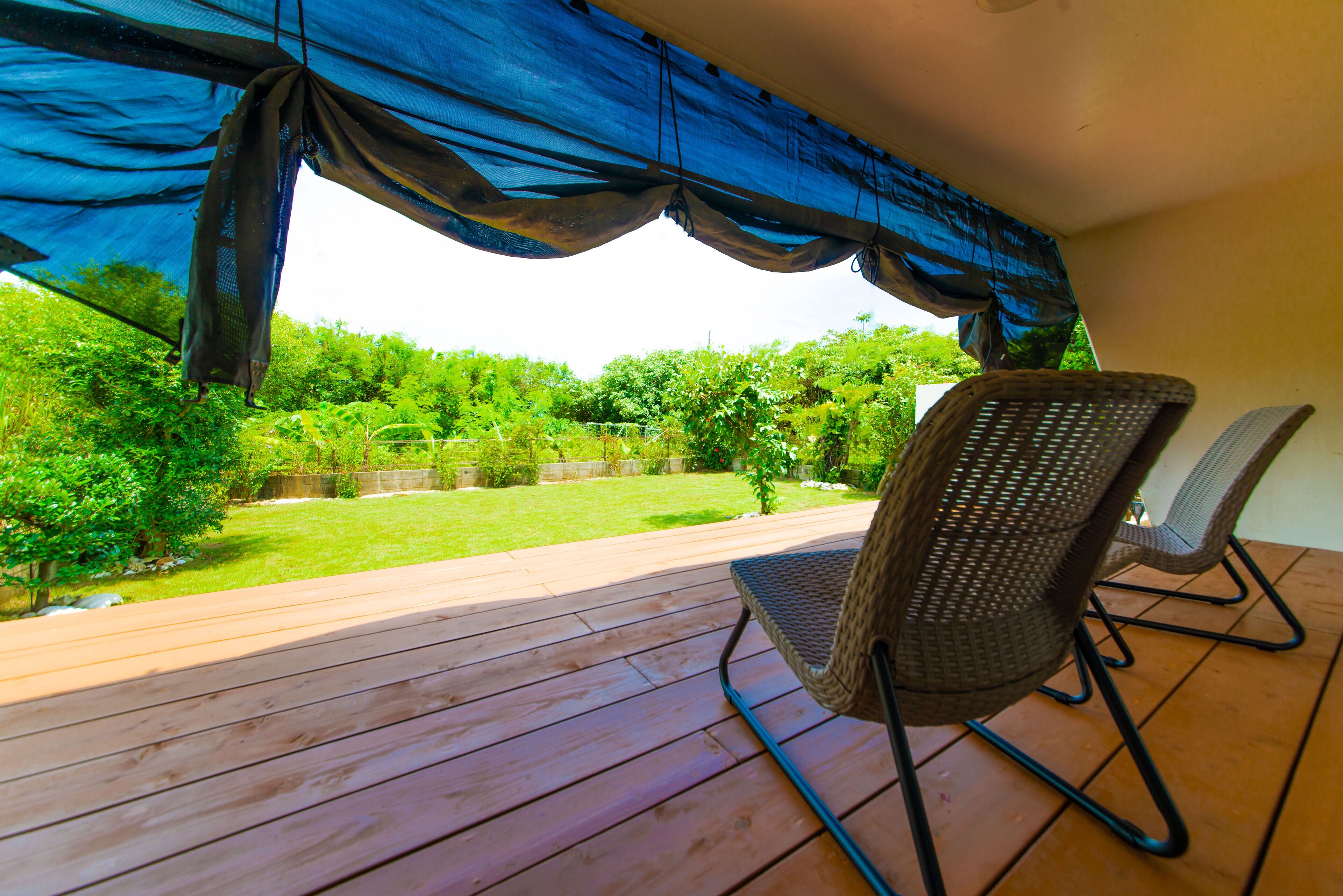 A covered outdoor area with a wooden deck, wicker chairs, and a view of a green garden.