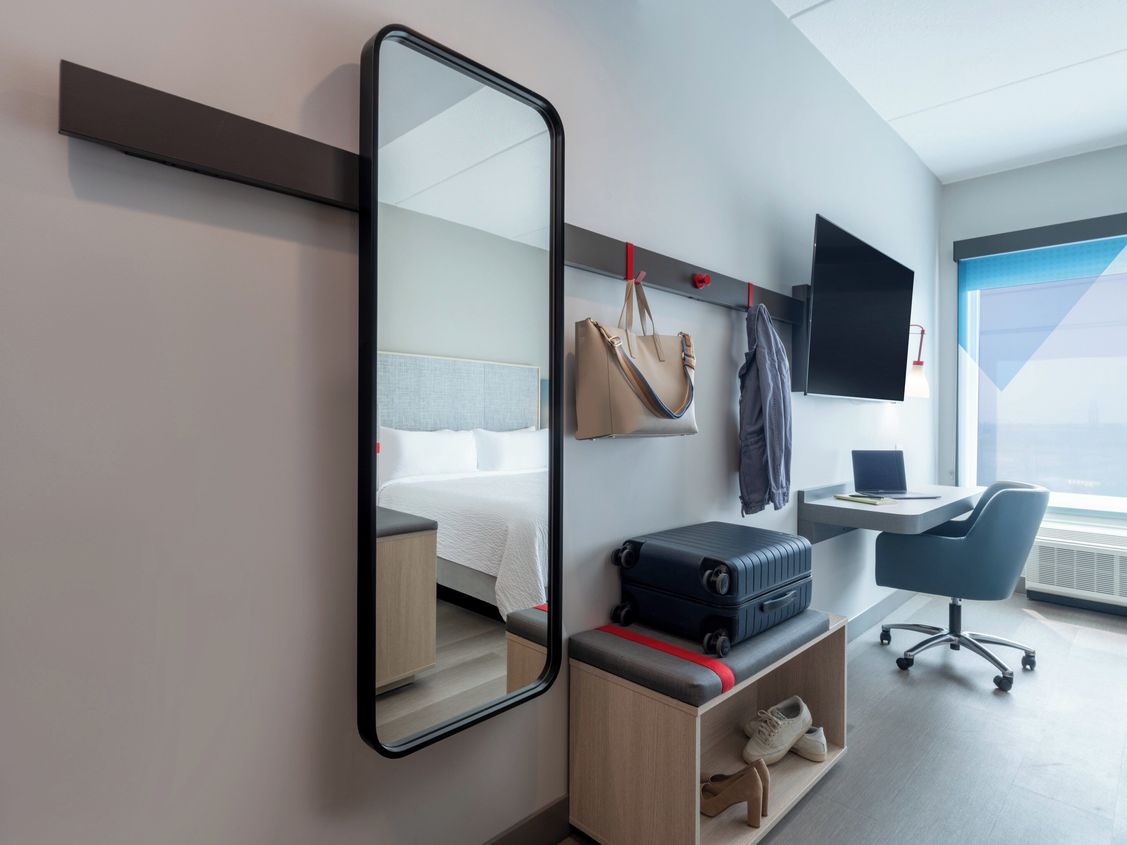 A modern hotel room with a bed, a desk with a laptop, a chair, a large mirror, and a wardrobe.