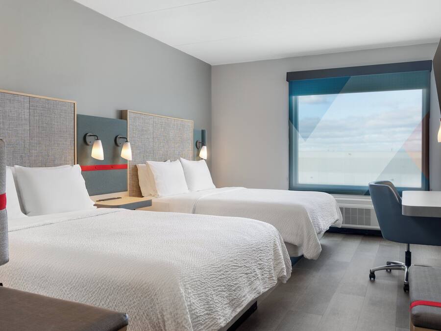 A hotel room with two beds, a desk, a chair, and a large window offering a view.