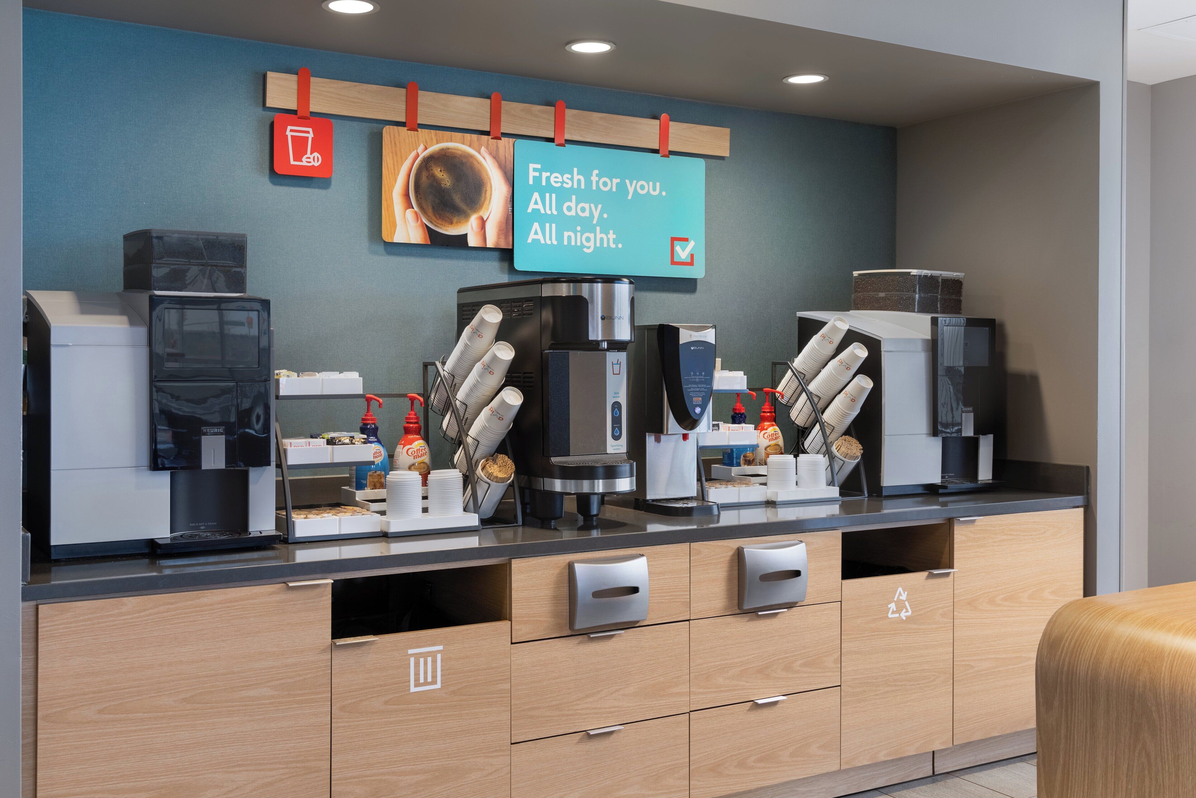 A modern coffee station with multiple coffee machines, a sign that reads "Fresh for you. All day. All night.", and a recycling bin.