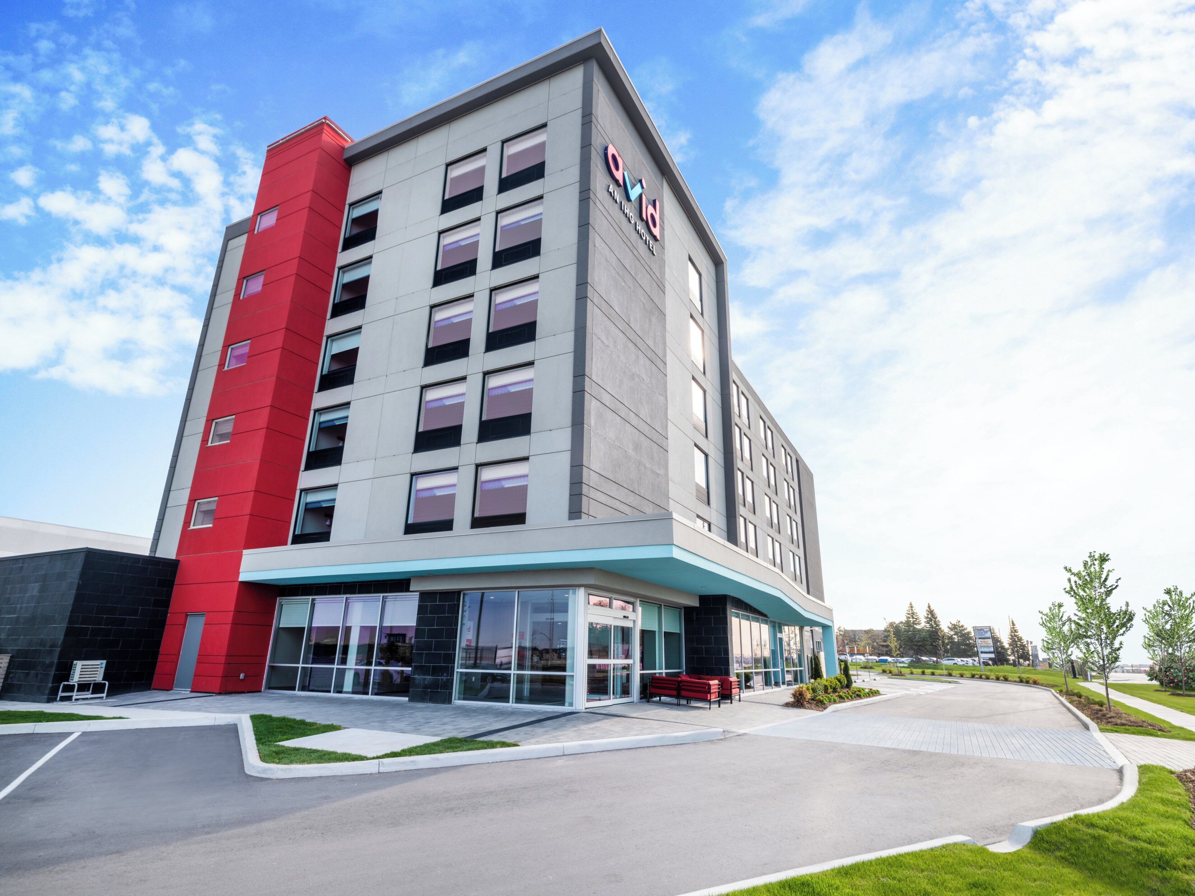 A modern hotel building with a prominent red and grey facade, large windows, and a well-maintained outdoor area with seating.