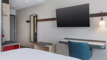 A modern hotel room with a large bed, a desk with a chair, a mounted TV, and a bedside table.