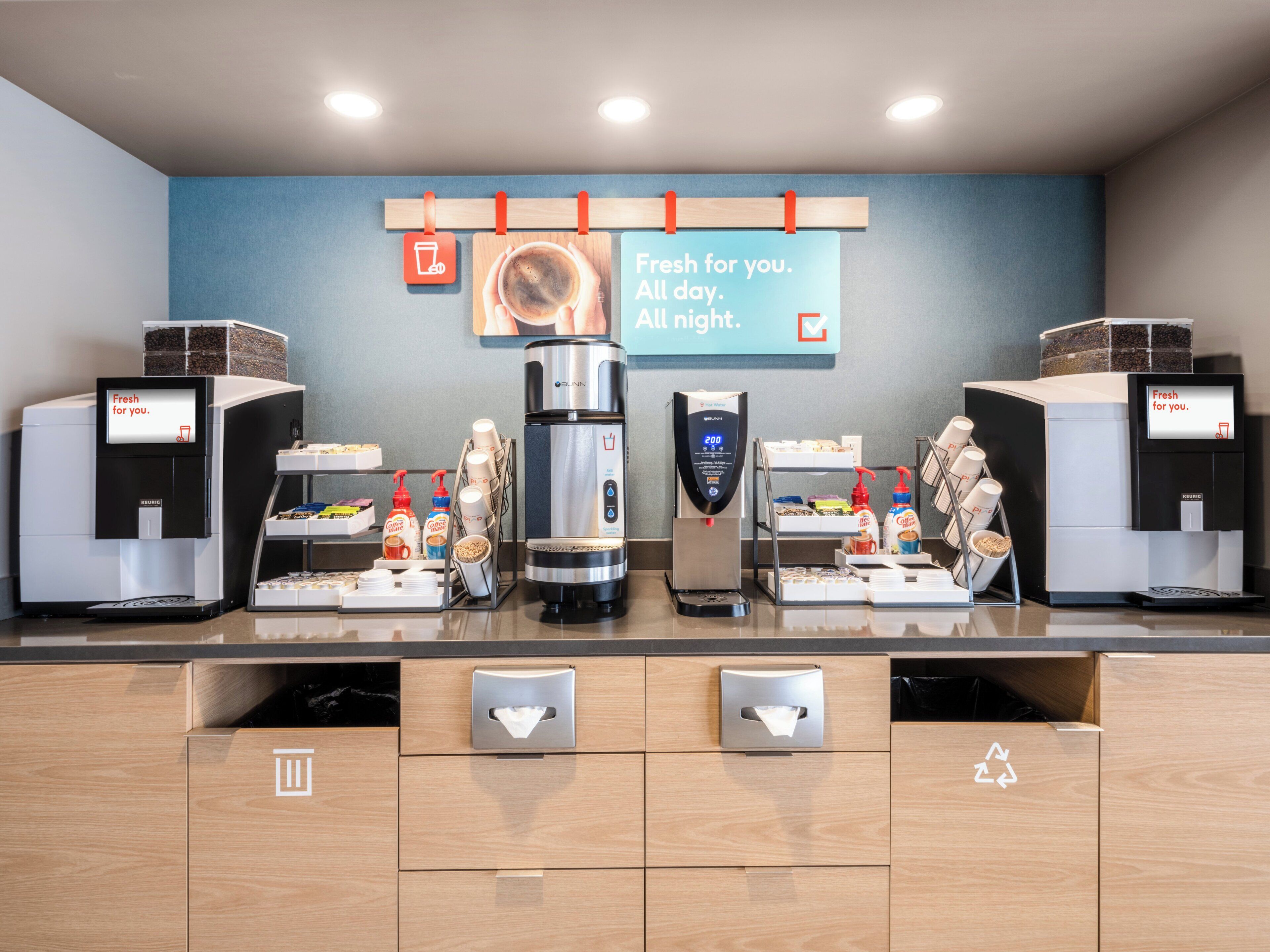 A coffee station with multiple coffee machines, a sign indicating availability of coffee all day and night, and a recycling bin.