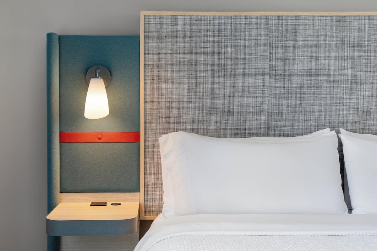 Quarto de hotel com cama, mesa de cabeceira, luminária fixada na parede e cabeceira texturizada.