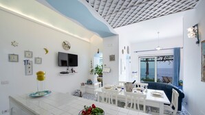 A modern kitchen with white tiles, a dining area with white chairs, a TV mounted on the wall, and a view of the sea through a large window.