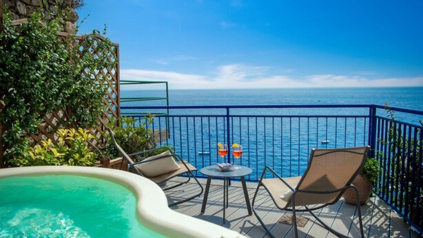 A balcony with a view of the sea, a hot tub, and two chairs with a table.