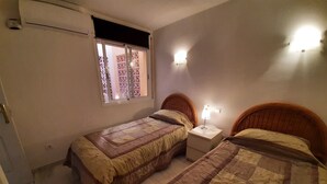 Een slaapkamer met twee eenpersoonsbedden, een nachtkastje met een lamp, een raam met uitzicht en een airconditioner.