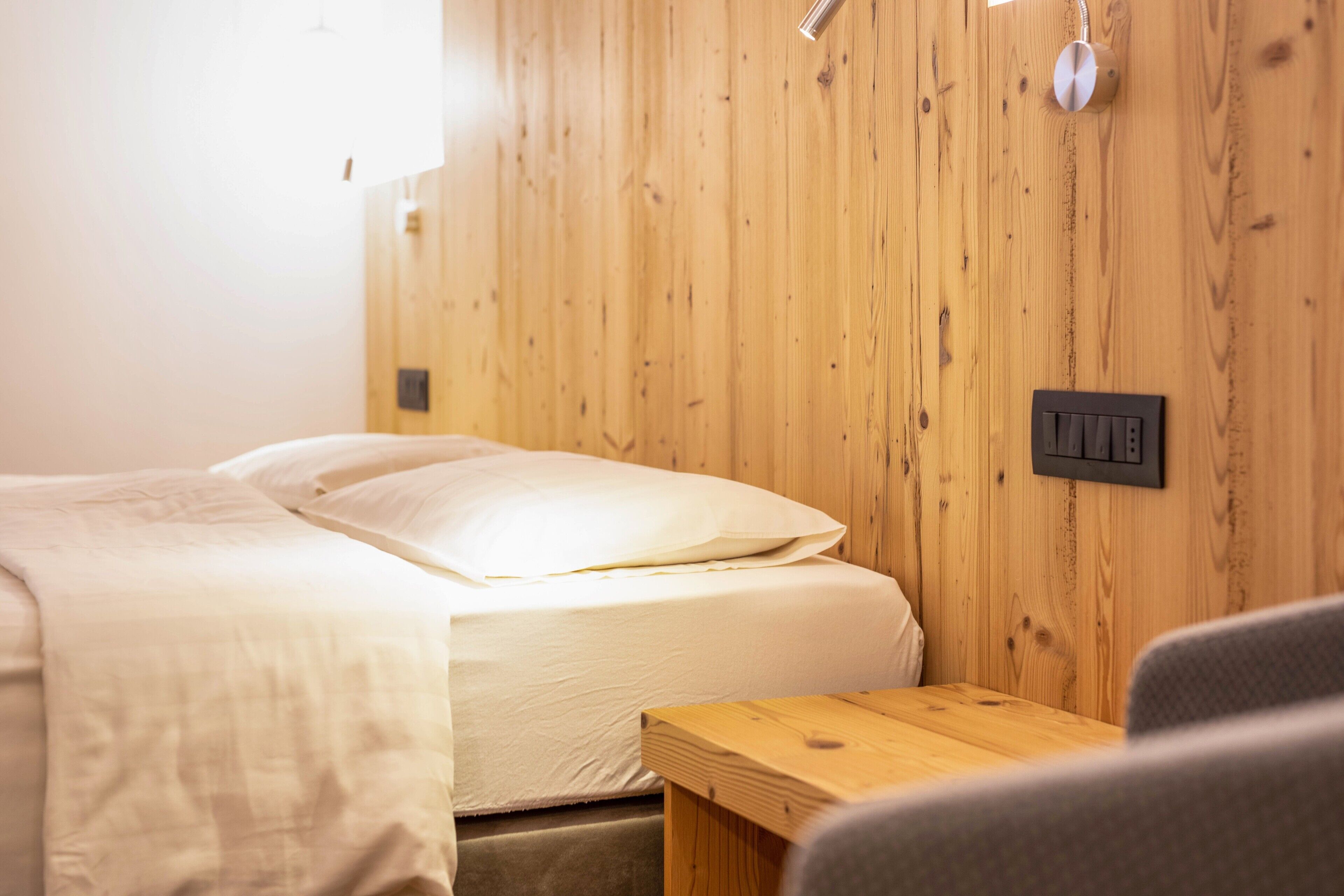 A room with a wooden wall, a bed with white bedding, a small table, and a wall-mounted light switch.